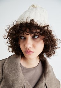 Beanie a coste crema con pompon, indossato da una persona con capelli ricci, abbinato a un cappotto marrone e una maglia grigia. Primo piano del viso.