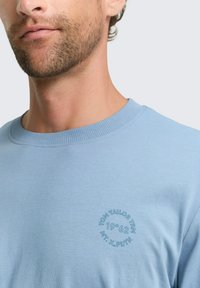 Hellblaues Baumwoll-T-Shirt mit einem gerippten Rundhalsausschnitt und einem kreisförmigen Logodetail in dunklerem Blau im linken Brustbereich.
