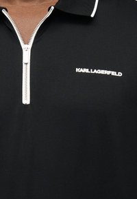 Μαύρο πουκάμισο με φερμουάρ στον γιακά, λευκό φερμουάρ και λευκή επιγραφή "KARL LAGERFELD" στο στήθος, φορεμένο από άτομο με μεσαία καστανή επιδερμίδα.