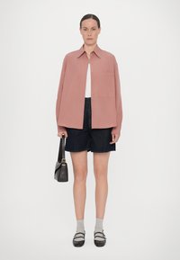 Chemise rose surdimensionnée avec un col, associée à un short en denim foncé. Le modèle tient un sac à bandoulière noir. Des chaussettes grises et des chaussures noires complètent le look.