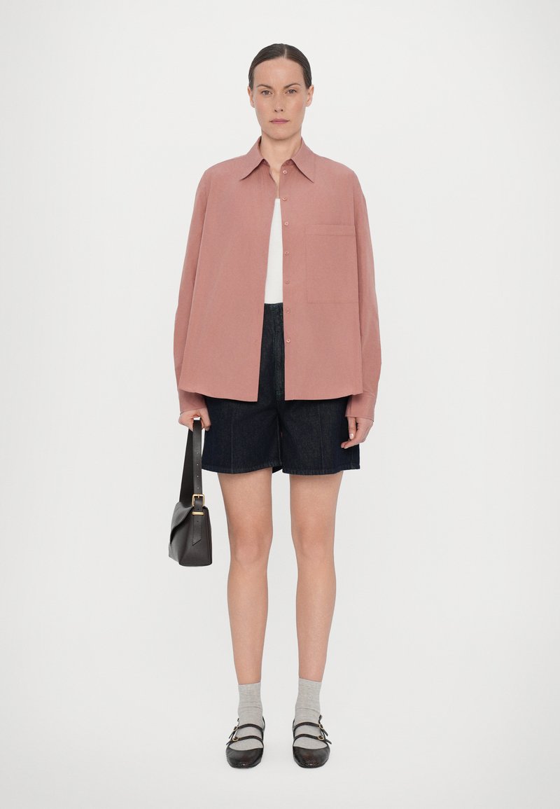 Rosa übergroßes Hemd mit Kragen, kombiniert mit dunklen Jeans-Shorts. Das Model hält eine schwarze Schultertasche. Graue Socken und dunkle Schuhe runden den Look ab.