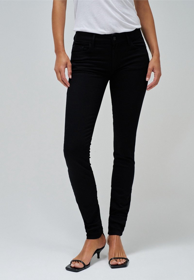 Femme portant un jean noir ajusté, une chemise blanche rentrée, et des sandales noires à talons avec lanières sur un fond clair uni.