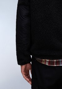 Giacca in pile nera con finitura testurizzata, maniche in nylon nero liscio a contrasto e un'emulsione di camicia a quadri visibile sotto.