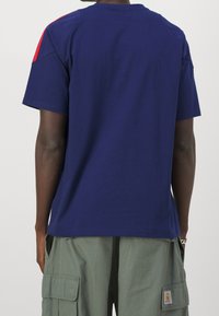 Granatowa bawełniana koszulka z krótkim rękawem z czerwonymi akcentami na ramionach, widoczna z tyłu, o luźnym kroju.