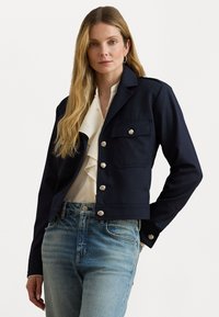 Donna con lunghi capelli biondi indossa una giacca blu navy con bottoni sopra una camicetta bianca con volant e jeans blu sbiaditi, in piedi di fronte a uno sfondo semplice.