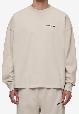 Beżowy, oversize sweatshirt z okrągłym dekoltem i opadającymi ramionami. Na klatce piersiowej znajduje się czarny napis "PEGADOR". Wykonany z miękkiego bawełnianego materiału.
