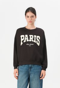 Woman wearing black sweatshirt with "PARIS Avec Amour 1986" text and blue jeans, standing against a plain white background.