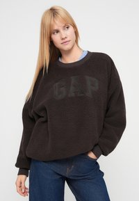 Sudadera tipo pulóver de forro polar marrón oscuro con "GAP" impreso en negro, puños acanalados y corte holgado. Se lleva sobre una camisa de rayas y combinada con vaqueros.