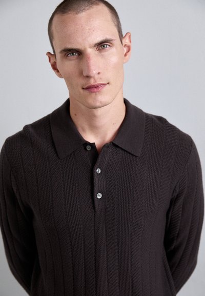 Mlad moški s kratkimi lasmi, oblečen v temno rjavo strukturirano pleteno polo majico, stoji pred enotno svetlo sivo ozadje.