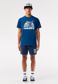 Camiseta navy con un juguetón logo de Lacoste y gráficos retro, combinada con pantalones cortos a juego, zapatillas azules y una gorra navy con el nombre de la marca.