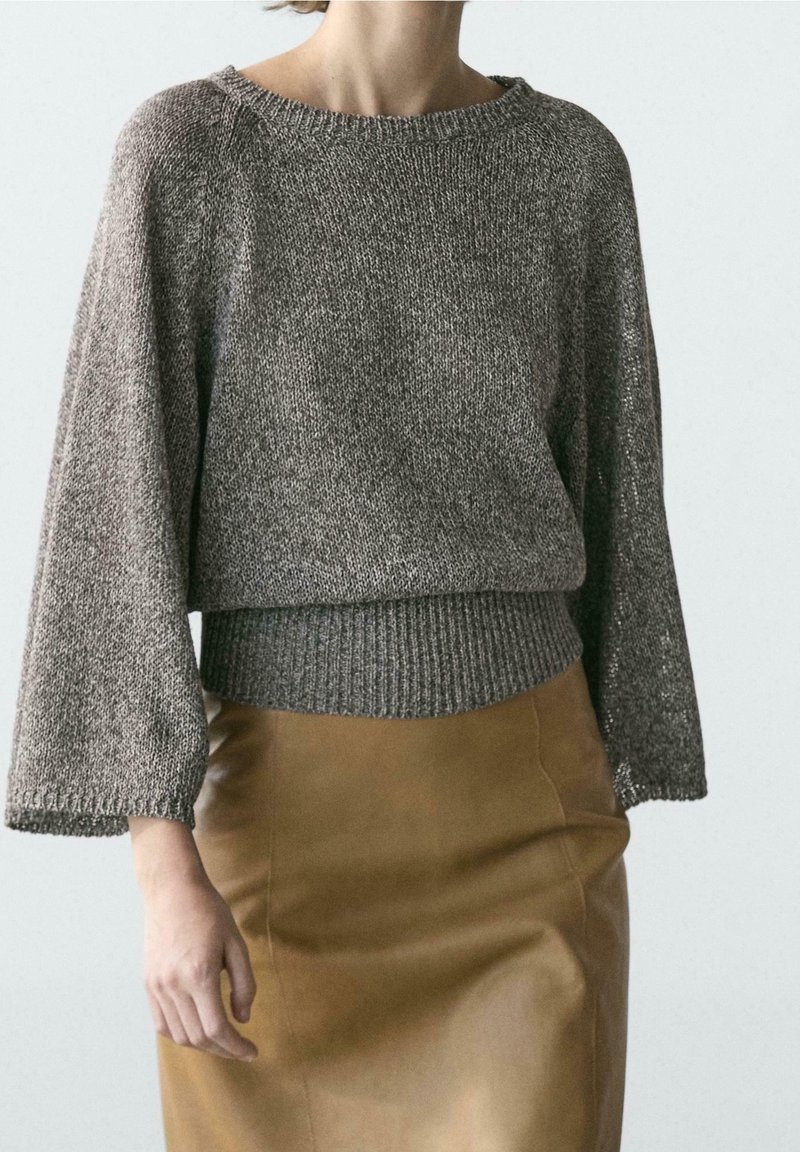 Personne portant un pull en maille gris ample avec de larges manches et une jupe en cuir marron cintrée sur un fond clair uni.