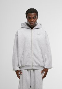 Felpa grigia con zip realizzata in tessuto morbido e dal taglio comodo. Presenta un cappuccio, tasche frontali e un ricamo logo discreto sul petto.