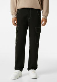 Black cargo pants with contrasting white stitching, multiple side pockets, and a relaxed fit, paired with white sneakers.