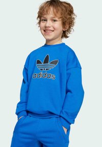 Sweatshirt bleu à col rond, au design surdimensionné, avec le logo Adidas noir. Texture douce, coupe décontractée, associé à un pantalon bleu assorti.