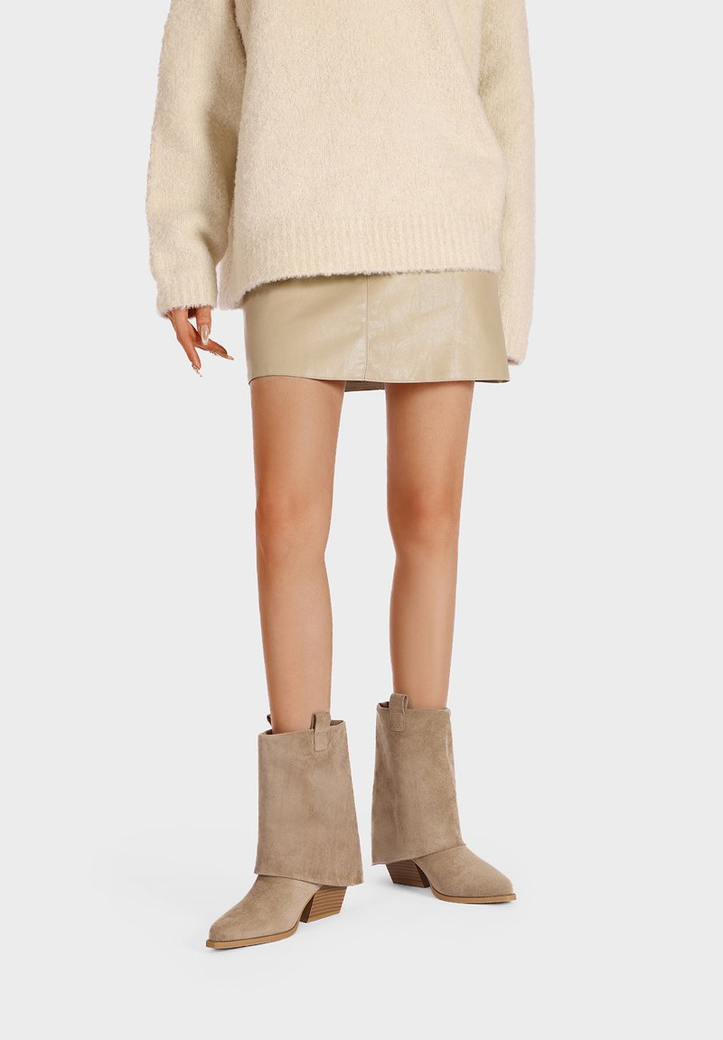 Stivaletti alla caviglia in suede beige con tacco spesso, caratterizzati da una punta squadrata e anelli laterali per una facile vestibilità. Abbinati a una minigonna beige chiaro.