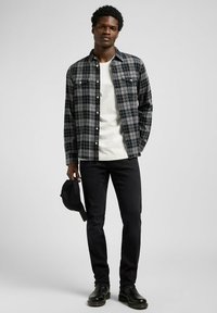 Męska koszula flanelowa w kratę w kolorach czarnym, szarym i białym, zapinana na guziki z dwoma kieszeniami na piersi, zestawiona z białym t-shirtem i czarnymi dżinsami. Trzymana czarna czapka.