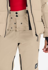 Beige pufferjacka med svarta dragkedjor, bärs över tan skidbyxor med tryckknappar och dragkedjefickor. Röd topp under.