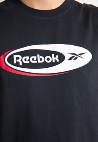 Juodas marškinėlis su dideliu baltu ir raudonu Reebok logotipu. Audinys atrodo lygus, su klasikiniu apvaliu kaklu.
