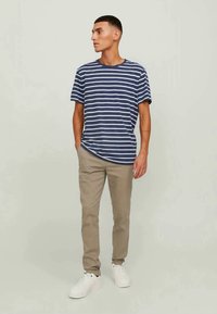 Homem a usar uma t-shirt riscada em azul e branco, calças bege e ténis brancos, de pé com as mãos nos bolsos, a olhar para o lado.