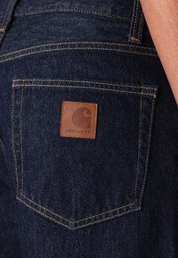 Jeans de mezclilla azul oscuro con un parche de marca de cuero marrón en el bolsillo trasero, con costuras amarillas contrastantes y un corte clásico.