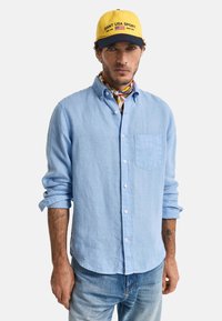 Camisa de lino azul claro con mangas largas, frente abotonado, un bolsillo en el pecho y puños enrollados. Combinada con una gorra amarilla y una bufanda de cuello colorida.