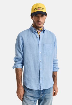 Chemise en lin bleu clair avec des manches longues, devant boutonné, poche poitrine unique et poignets retroussés. Portée avec une casquette jaune et un foulard coloré autour du cou.