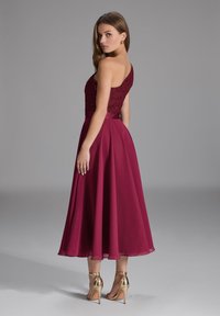Burgundy -värinen mekko, jonka yläosassa on teksturoitu pitsi ja alhaalla virtaava sifonkihame. Mekossa on yksittäinen olkaimellinen muotoilu, ja se on yhdistetty kultaisiin korkokenkiin.