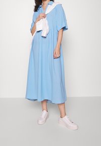 Ljusblå randig klänning med korta ärmar och krage; har en insydd midja. Bärs med vita sneakers, visad mot en neutral bakgrund.