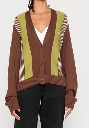 Kvinde iført brun cardigan med knapper, grønne og hvide lodrette striber samt sorte bukser, hænder afslappet langs siden.