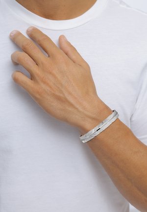 Silver metal bracelet with a textured, layered design, worn on a hand against a white shirt background. Smooth finish, slim profile.