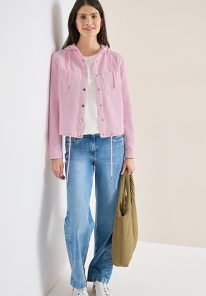 Žena v růžové bundě, bílém tričku, modrých džínách a bílých teniskách, držící olivově zelenou tašku, stojící před dvoubarevnou béžovou zdí.