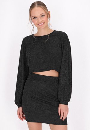 Jonge vrouw lacht terwijl ze een zwart cropped top met lange mouwen en een bijpassende zwarte rok draagt tegen een witte achtergrond.
