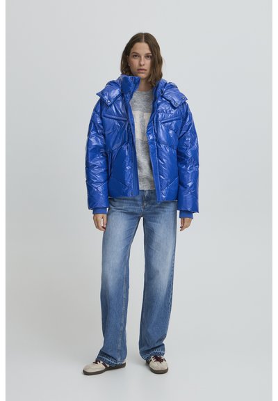 Kék puffertakaru dzseki fényes felülettel, magas gallérral és nagy zsebekkel. Szürke pulóver fölött viseli, világos farmernadrággal párosítva.