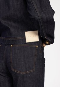 Donkerdenim jeans hebben een leren label met "AAIKO" op de achterkant, oranje stiksels en een klassiek zakontwerp met gouden hardwareaccenten.