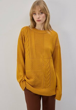 Femme blonde portant un pull en maille texturée jaune moutarde et un pantalon marron, debout contre un mur gris clair uni.