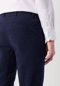 Närbild av en person som bär marinblå Calvin Klein-byxor och en vit knappad skjorta, som visar bakfickan och handen.