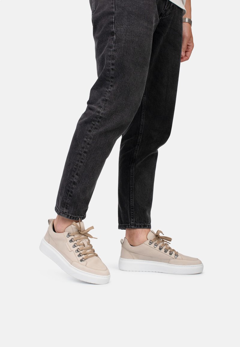 Beige leren sneakers met ronde neus en witte rubberen zool. Voorzien van metalen veterogen en tan veters. Gecombineerd met donkere spijkerbroek.