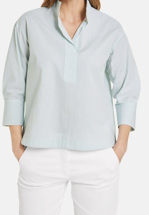 Femme portant une chemise à manches longues rayée bleu clair avec des poignets repliés et un pantalon blanc, les mains dans les poches, sur un fond blanc.
