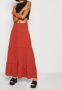 Jupe maxi à volants de couleur rouge, fabriquée en tissu doux, dotée d'une taille élastique et d'un ourlet à volants, associée à des sandales à plateforme noires.