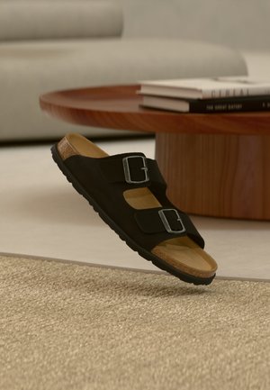 Sandale noire à deux lanières avec boucles argentées et semelle en liège reposant sur un tapis beige, table basse ronde en bois et livres en arrière-plan.