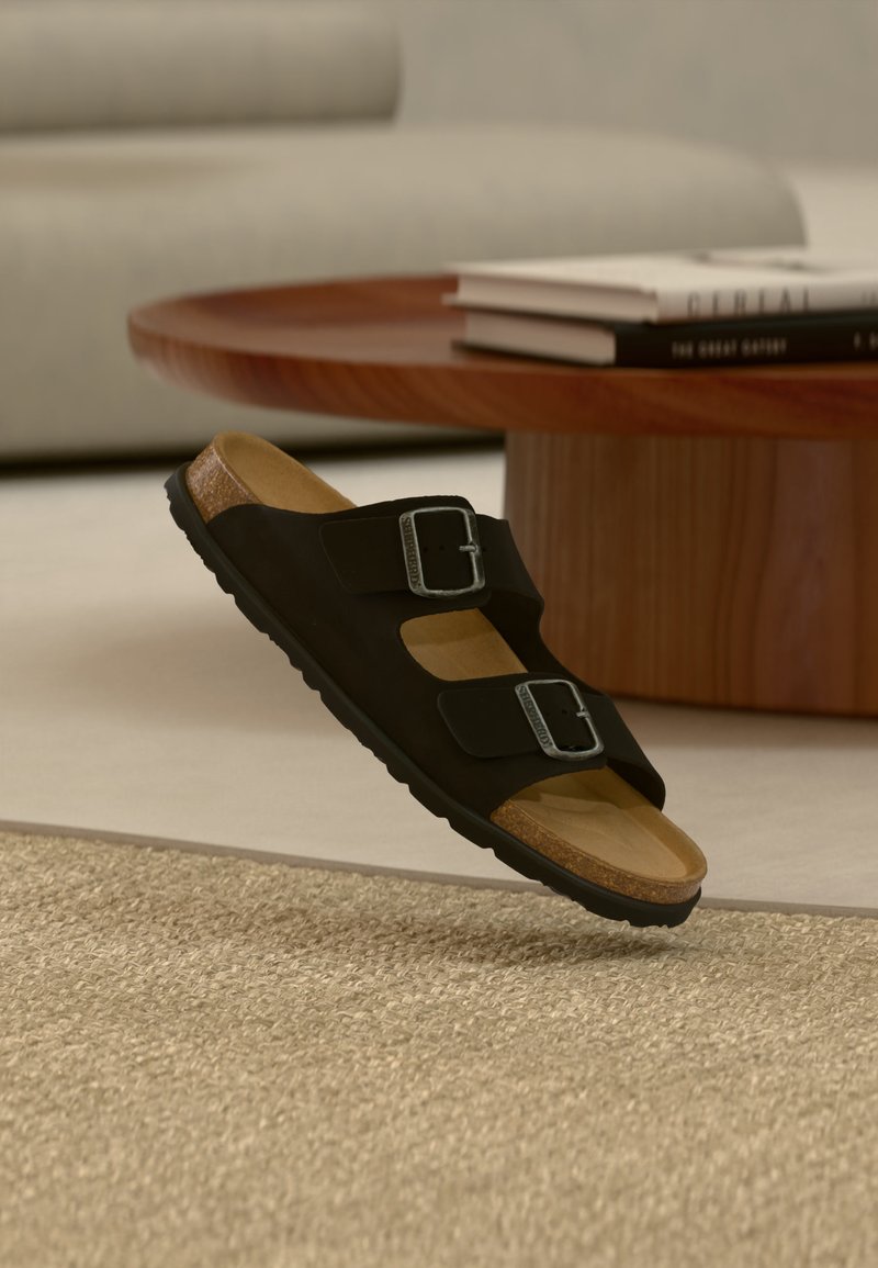 Μαύρο σανδάλι με δύο λουριά και ασημί αγκράφες, με πάτο από φελλό, ακουμπισμένο σε μπεζ χαλί, με ξύλινο στρογγυλό τραπεζάκι καφέ και βιβλία στο φόντο.