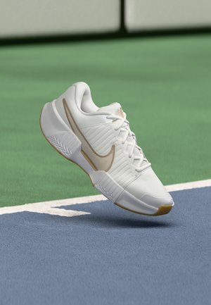 Baskets Nike blanches et beiges suspendues au-dessus d’une surface de court de tennis bleu et vert près des lignes blanches de délimitation.