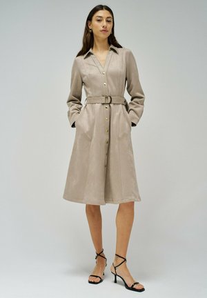 Robe en daim beige avec un col, boutonnière à l'avant, taille ceinturée et poches latérales. Dotée de manches longues et d'un ourlet évasé aux genoux.