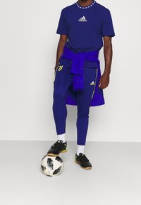 Niebieska koszulka Adidas z logo, pasujące spodnie dresowe z żółtymi akcentami i czarne buty piłkarskie na piłce nożnej.