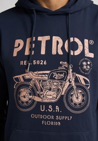 Donkerblauwe hoodie met een grote retro motorfietsafbeelding in het roze en de tekst "PETROL" met verouderde details. Zacht materiaal met een capuchon met trekkoord.