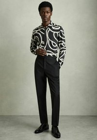 Camicia a righe in bianco e nero con disegni vorticosi, abbinata a pantaloni neri su misura e scarpe di pelle nere. Texture del tessuto liscia.