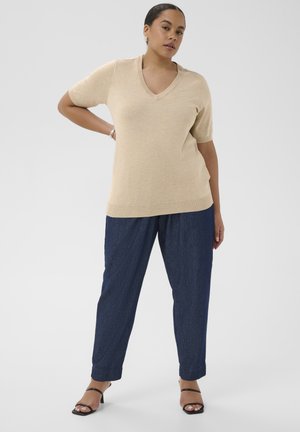 Femme portant un pull beige à manches courtes avec un col en V, un pantalon ample bleu foncé, des talons noirs à lanières, posant avec une main sur la hanche.