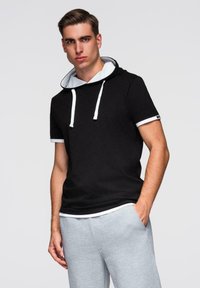 Mladý muž v černé mikině s krátkým rukávem a bílými tkaničkami, v šedých teplácích, stojící s jednou rukou v kapse na jednobarevném pozadí.