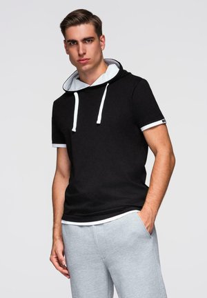 Jeune homme en hoodie noir à manches courtes avec cordons blancs et jogging gris, debout avec une main dans la poche devant un fond uni.