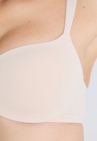 Κοντινό πλάνο ενός ατόμου που φοράει ένα λείο, ανοιχτό μπεζ σουτιέν με αδιάλειπτες θήκες και ρυθμιζόμενες τιράντες, σε ουδέτερο φόντο.